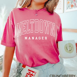 May include: A pink t-shirt with the text "MELTDOWN MANAGER" in white. The shirt is being worn by a person with a smile. The person is holding a book titled "How to Grow House Plants" and a white mug with a smiley face.