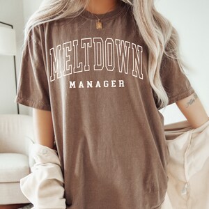 May include: A brown oversized t-shirt with the text "MELTDOWN MANAGER" printed in white.