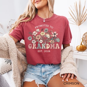 May include: Crimson-colored t-shirt with the text "PROMOTED TO GRANDMA EST. 2026" in a retro font. The shirt features a floral design in pastel colors. The shirt is short-sleeved and made of a soft material.