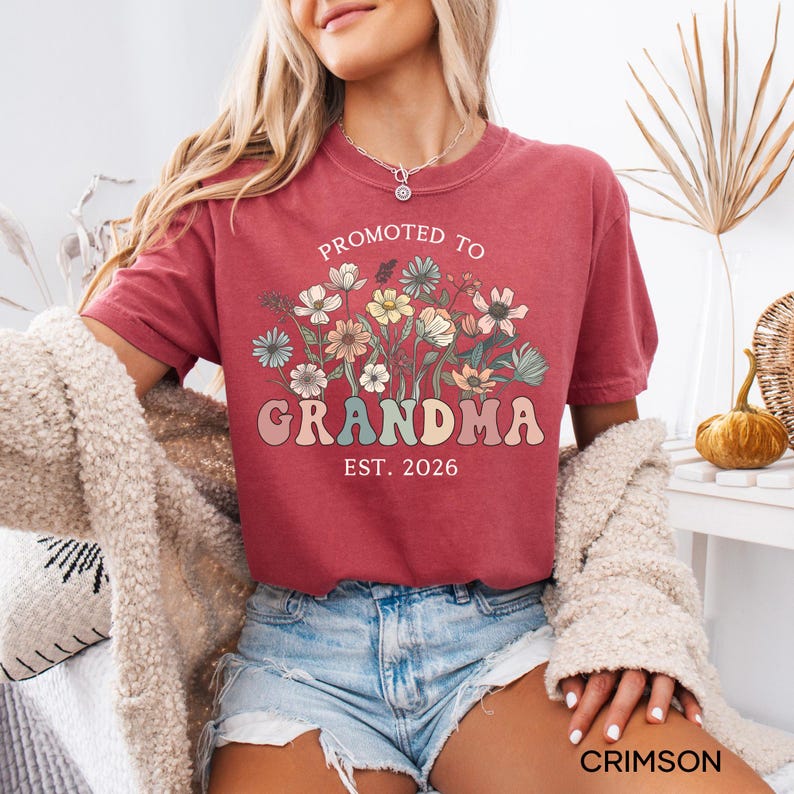 May include: Crimson-colored t-shirt with the text "PROMOTED TO GRANDMA EST. 2026" in a retro font. The shirt features a floral design in pastel colors. The shirt is short-sleeved and made of a soft material.