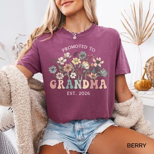 May include: Berry-colored t-shirt with the text "Promoted to Grandma Est. 2026" in a retro font, accompanied by a floral design. The shirt is short-sleeved and has a relaxed fit, perfect for casual wear.