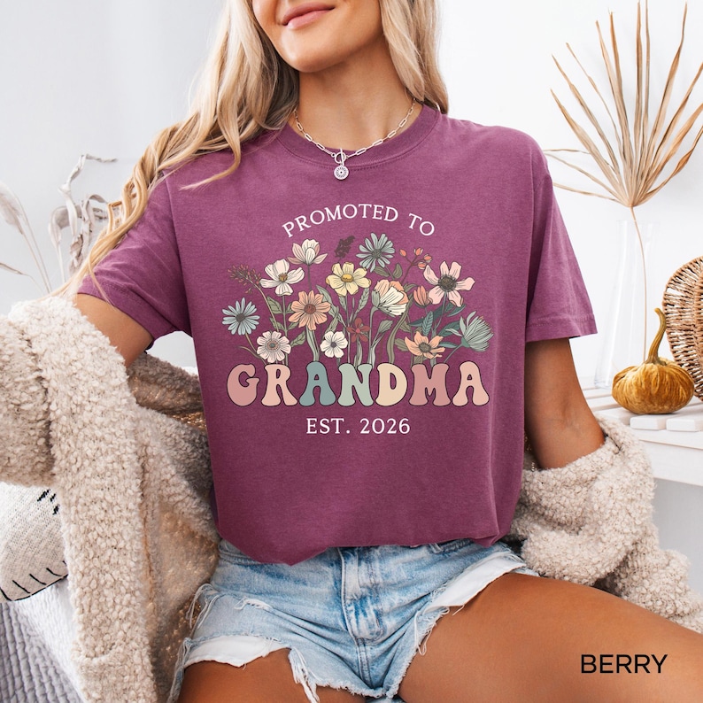 May include: Berry-colored t-shirt with the text "Promoted to Grandma Est. 2026" in a retro font, accompanied by a floral design. The shirt is short-sleeved and has a relaxed fit, perfect for casual wear.