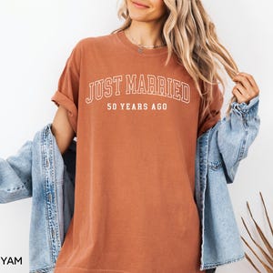 May include: A rust-colored t-shirt with the text "JUST MARRIED" in white, arched across the top, and "50 YEARS AGO" below. The shirt is worn with a light blue denim jacket.