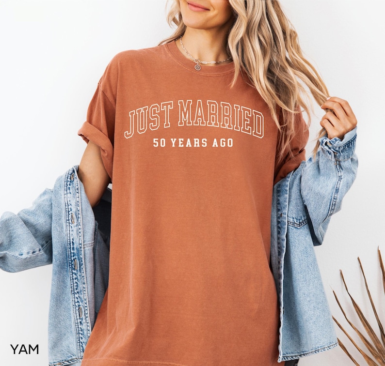 May include: A rust-colored t-shirt with the text "JUST MARRIED" in white, arched across the top, and "50 YEARS AGO" below. The shirt is worn with a light blue denim jacket.
