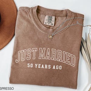 May include: Espresso-colored t-shirt with the words "JUST MARRIED" and "50 YEARS AGO" in white block letters. A silver necklace with a pendant rests on the shirt. The shirt is folded and has a Comfort Colors tag.