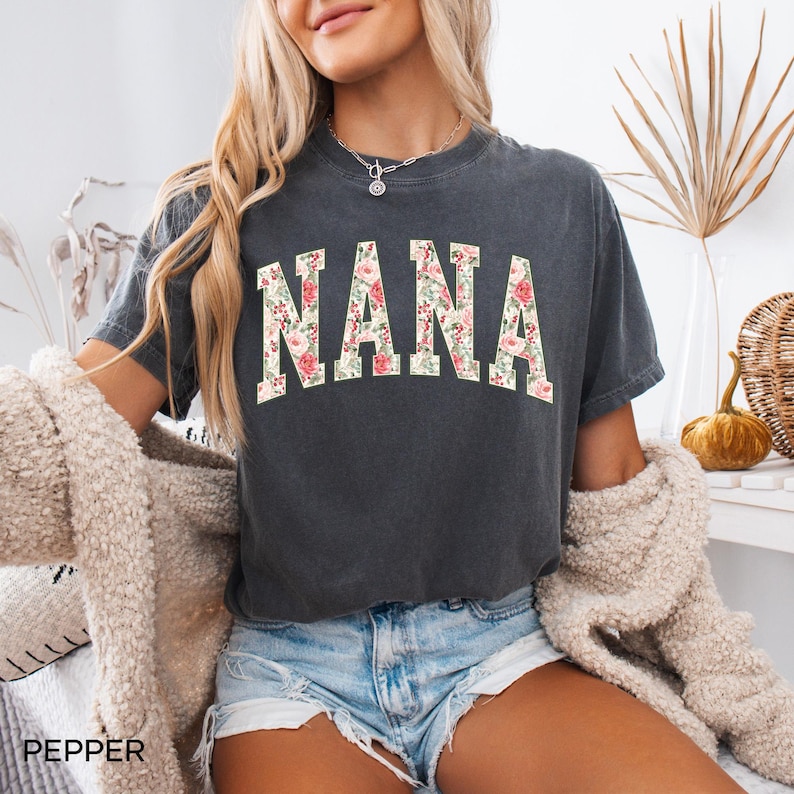 May include: Dark gray t-shirt with the word "NANA" in a floral design. The shirt is short-sleeved and has a vintage, worn look. The word "PEPPER" is printed at the bottom left of the image.