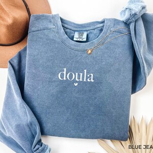 May include: A blue jean-coloured sweatshirt with the word "doula" embroidered in white, with a small heart beneath. The sweatshirt has a crew neck and long sleeves. A gold necklace with a pendant is draped across the front. The tag reads "Comfort Colors".
