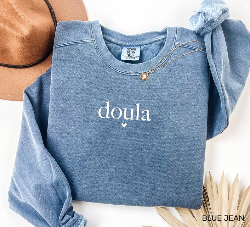 May include: A blue jean-coloured sweatshirt with the word "doula" embroidered in white, with a small heart beneath. The sweatshirt has a crew neck and long sleeves. A gold necklace with a pendant is draped across the front. The tag reads "Comfort Colors".