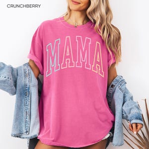 May include: A pink t-shirt with the word "MAMA" in rainbow colors. The shirt is a relaxed fit and has a crew neck.