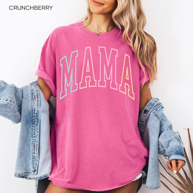 May include: A pink t-shirt with the word "MAMA" in rainbow colors. The shirt is a relaxed fit and has a crew neck.