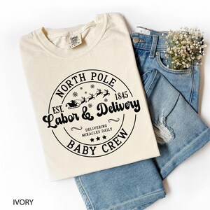 May include: Ivory-colored t-shirt with a black graphic that reads "North Pole Labor & Delivery Baby Crew" with a Santa sleigh design. The shirt is paired with light blue denim jeans and a small bouquet of white flowers.