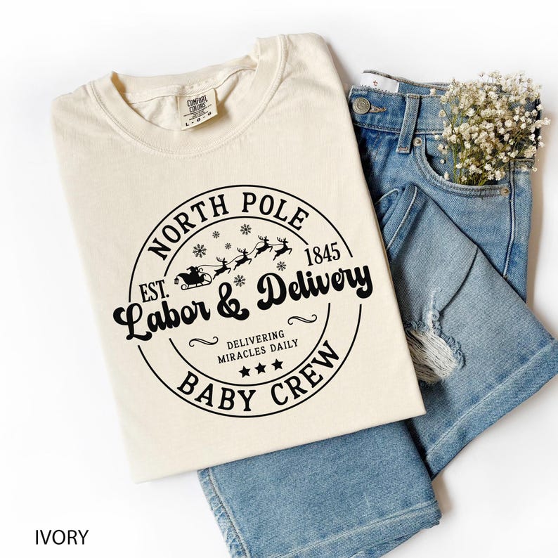 May include: Ivory-colored t-shirt with a black graphic that reads "North Pole Labor & Delivery Baby Crew" with a Santa sleigh design. The shirt is paired with light blue denim jeans and a small bouquet of white flowers.