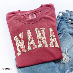 May include: Crimson-colored Comfort Colors t-shirt with the word "NANA" in large, floral-patterned letters. The shirt is folded next to a pair of blue denim jeans. The word "CRIMSON" is printed at the bottom left.