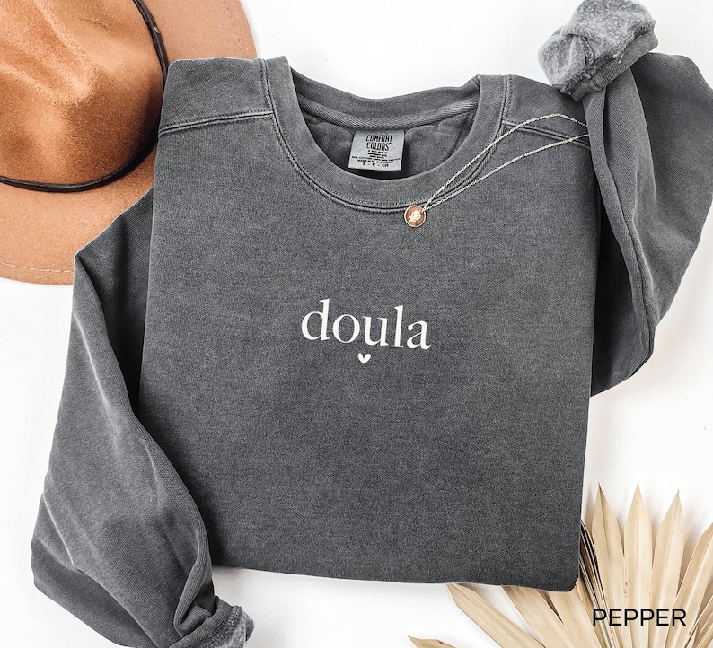 May include: A dark grey sweatshirt with the word "doula" in white script, with a small heart below. The sweatshirt has long sleeves and a crew neck. A gold necklace is draped across the front. A brown hat is in the background.