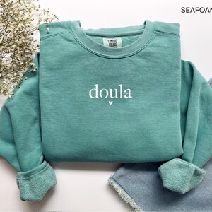 May include: Seafoam green sweatshirt featuring the word "doula" in white script, with a small heart beneath. The sweatshirt has a crew neck and ribbed cuffs. The brand "Comfort Colors" is visible on the tag.