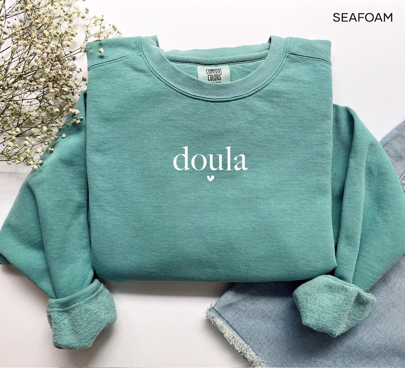 May include: Seafoam green sweatshirt featuring the word "doula" in white script, with a small heart beneath. The sweatshirt has a crew neck and ribbed cuffs. The brand "Comfort Colors" is visible on the tag.