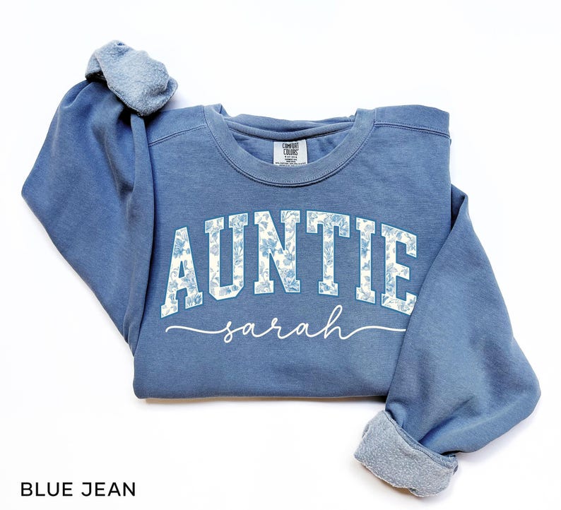 May include: Blue jean colored sweatshirt with the word "AUNTIE" in white block letters with a floral pattern, and the name "sarah" in cursive. The sweatshirt has a crew neck and long sleeves with ribbed cuffs.