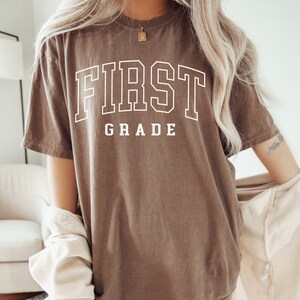 May include: A brown t-shirt with the text "FIRST GRADE" in white.