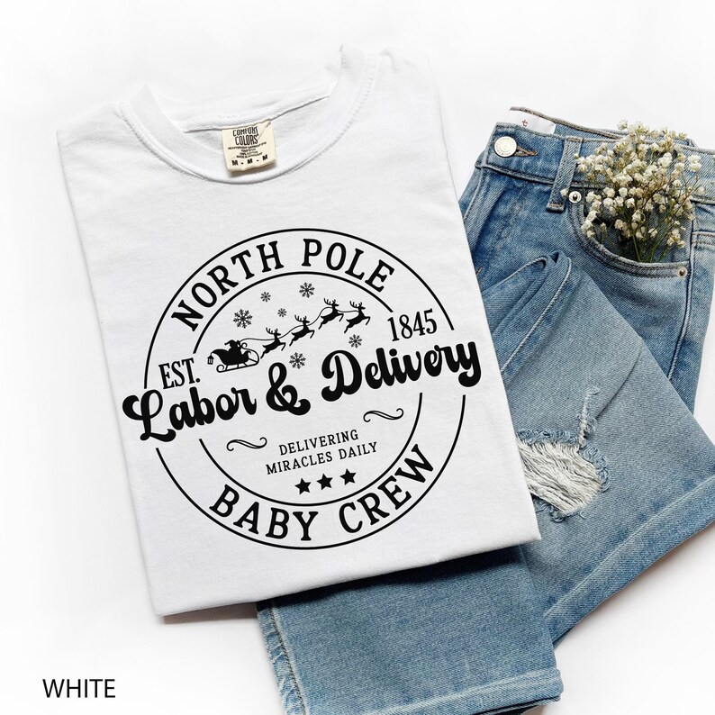 May include: White t-shirt with a round graphic that reads "NORTH POLE LABOR & DELIVERY BABY CREW" with a Santa sleigh and reindeer design. The shirt is paired with light blue denim jeans.