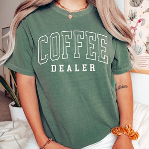 May include: Moss green t-shirt with the words "COFFEE DEALER" in white block letters. The shirt is a short-sleeved, crew neck style. The word "MOSS" is printed in the bottom left corner of the image.