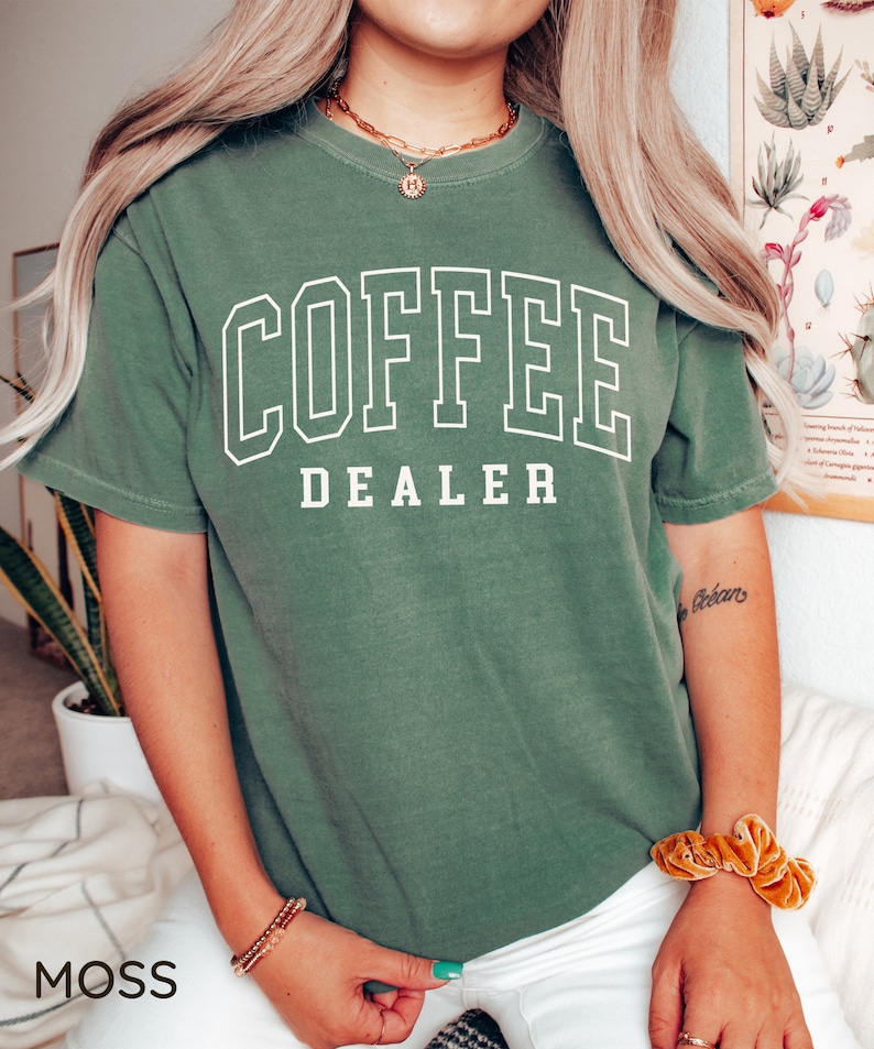May include: Moss green t-shirt with the words "COFFEE DEALER" in white block letters. The shirt is a short-sleeved, crew neck style. The word "MOSS" is printed in the bottom left corner of the image.