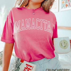 K&ouml;nnte beinhalten: Pinkes T-Shirt mit dem Wort "MAMACITA" in wei&szlig; auf der Vorderseite. Das Shirt wird von einer Person mit blonden Haaren und einem L&auml;cheln getragen. Die Person h&auml;lt ein Buch mit dem Titel "How to Grow House Plants" und eine wei&szlig;e Tasse mit einem Smiley.