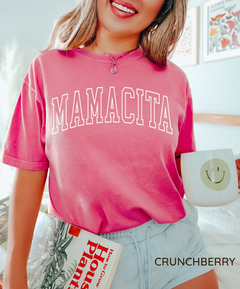 K&ouml;nnte beinhalten: Pinkes T-Shirt mit dem Wort "MAMACITA" in wei&szlig; auf der Vorderseite. Das Shirt wird von einer Person mit blonden Haaren und einem L&auml;cheln getragen. Die Person h&auml;lt ein Buch mit dem Titel "How to Grow House Plants" und eine wei&szlig;e Tasse mit einem Smiley.