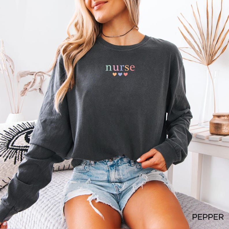 May include: A dark gray long-sleeve sweatshirt with the word "nurse" embroidered in pastel colors above three small hearts. The sweatshirt is paired with denim shorts. The word "PEPPER" is printed in the bottom right corner.