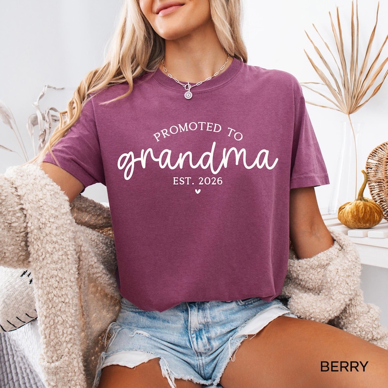 May include: Berry-colored t-shirt with the text "PROMOTED TO grandma EST. 2026" in white script. The shirt has a crew neck and short sleeves. The word "BERRY" is printed in the bottom right corner.