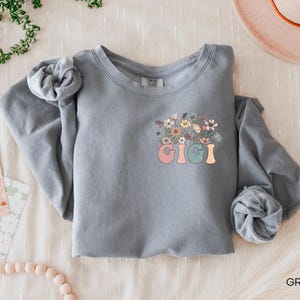 以下が含まれることがあります： 花柄のデザインとカラフルな文字で「Gigi」と書かれたグレーのスウェットシャツです。