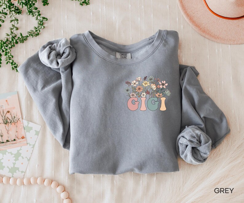 以下が含まれることがあります： 花柄のデザインとカラフルな文字で「Gigi」と書かれたグレーのスウェットシャツです。