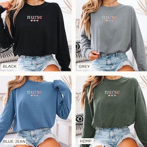 May include: Four long-sleeved shirts in black, grey, blue jean, and hemp colors. Each shirt has the word "nurse" embroidered in pastel colors above three hearts. The shirts are made of a soft material.