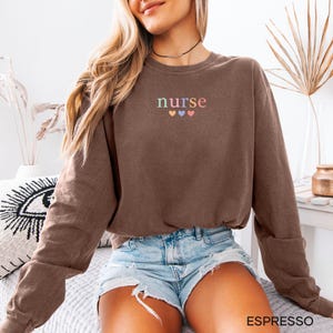 May include: Espresso-colored sweatshirt with the word "nurse" embroidered in pastel colors above three hearts. The long-sleeved crew neck top is paired with denim shorts. The word "ESPRESSO" is printed in a white font at the bottom of the image.