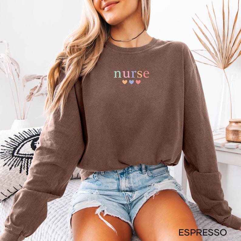 May include: Espresso-colored sweatshirt with the word "nurse" embroidered in pastel colors above three hearts. The long-sleeved crew neck top is paired with denim shorts. The word "ESPRESSO" is printed in a white font at the bottom of the image.