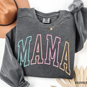 May include: A dark gray sweatshirt with the word "MAMA" printed in a rainbow color scheme. The letters are outlined in black and have a vintage collegiate style.