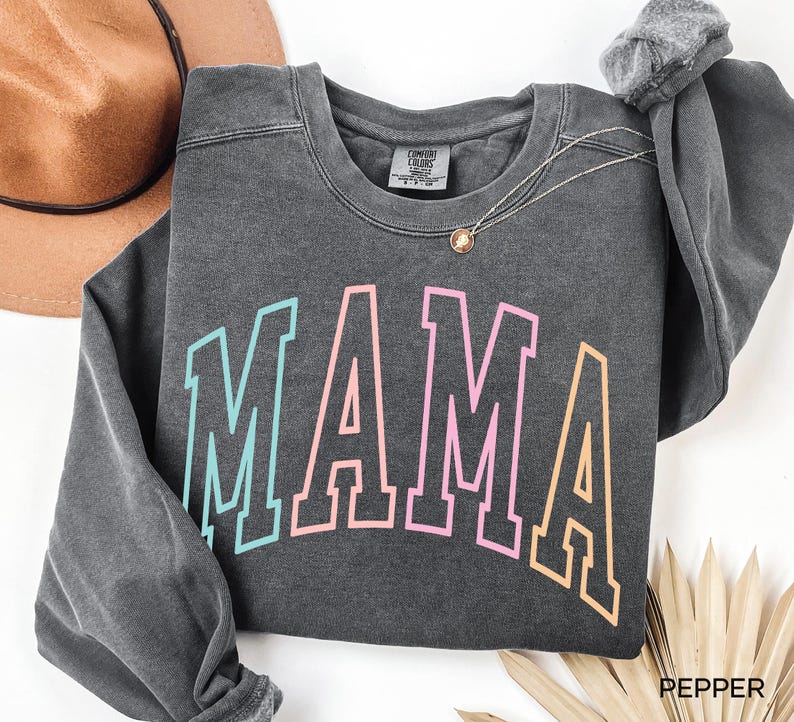 May include: A dark gray sweatshirt with the word "MAMA" printed in a rainbow color scheme. The letters are outlined in black and have a vintage collegiate style.