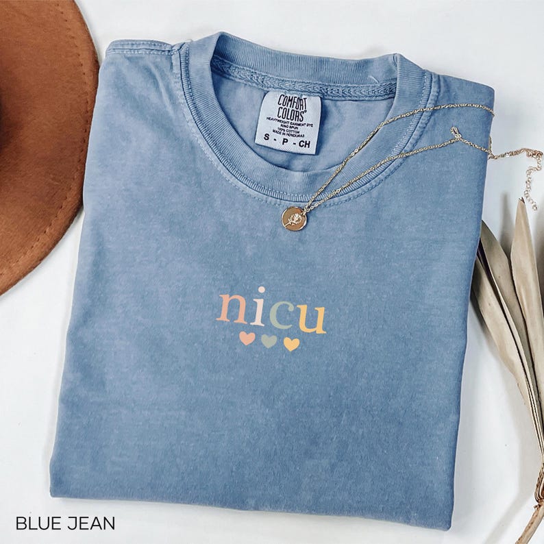 May include: A blue jean colored t-shirt with the word "nicu" in pastel colors and three hearts. A gold necklace with a pendant is draped across the shirt. The shirt is folded and has a tag that says "Comfort Colors".