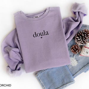 May include: A lilac-coloured sweatshirt with the word "doula" printed in black. The sweatshirt is folded on top of a pair of blue jeans, with a pinecone, a small gift bag, and a second pinecone. The sweatshirt has a soft, comfortable look.