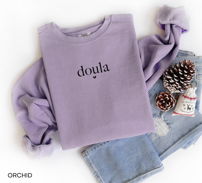 May include: A lilac-coloured sweatshirt with the word "doula" printed in black. The sweatshirt is folded on top of a pair of blue jeans, with a pinecone, a small gift bag, and a second pinecone. The sweatshirt has a soft, comfortable look.