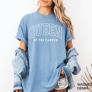 May include: A light blue t-shirt with the text "QUEEN OF THE CAMPER" in white outlined letters. The shirt is worn by a person with long blonde hair and a denim jacket.
