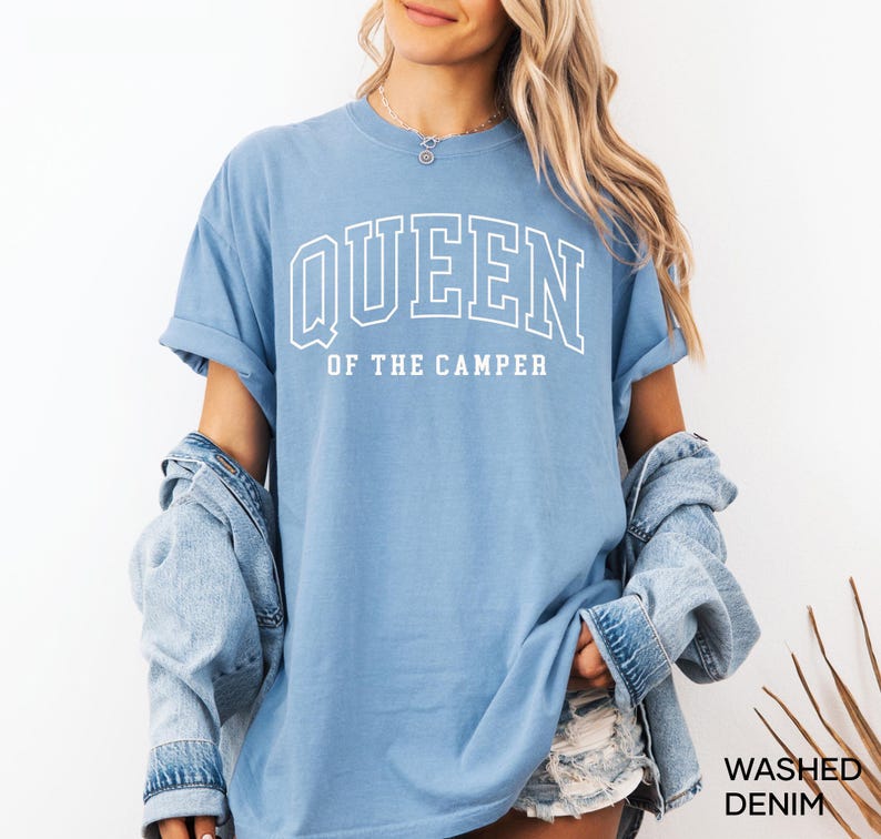 May include: A light blue t-shirt with the text "QUEEN OF THE CAMPER" in white outlined letters. The shirt is worn by a person with long blonde hair and a denim jacket.