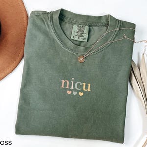 May include: A moss green t-shirt with the word "nicu" in pastel colors and three small hearts underneath. A gold necklace with a pendant rests on the shirt. The shirt is folded on a white surface.