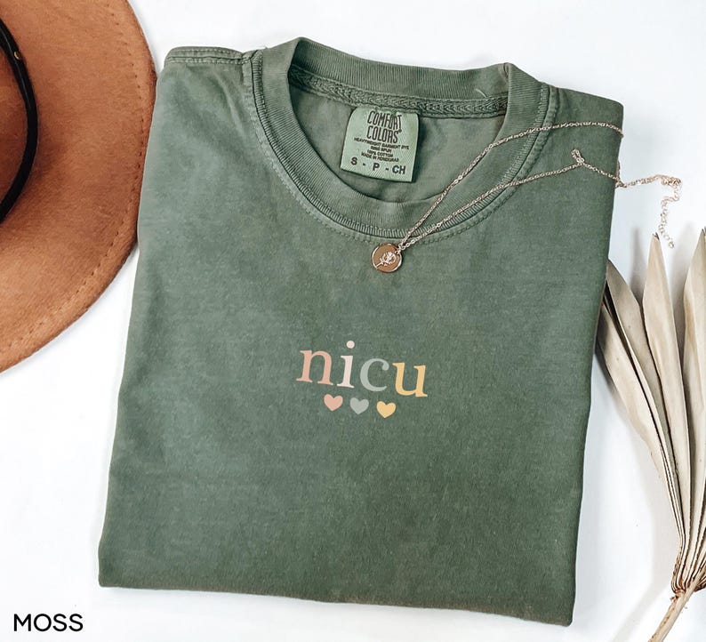 May include: A moss green t-shirt with the word "nicu" in pastel colors and three small hearts underneath. A gold necklace with a pendant rests on the shirt. The shirt is folded on a white surface.