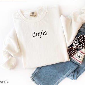 May include: White crew neck sweatshirt with the word "doula" printed in black. The sweatshirt is folded on top of blue jeans, with pinecones and a small decorative bag.