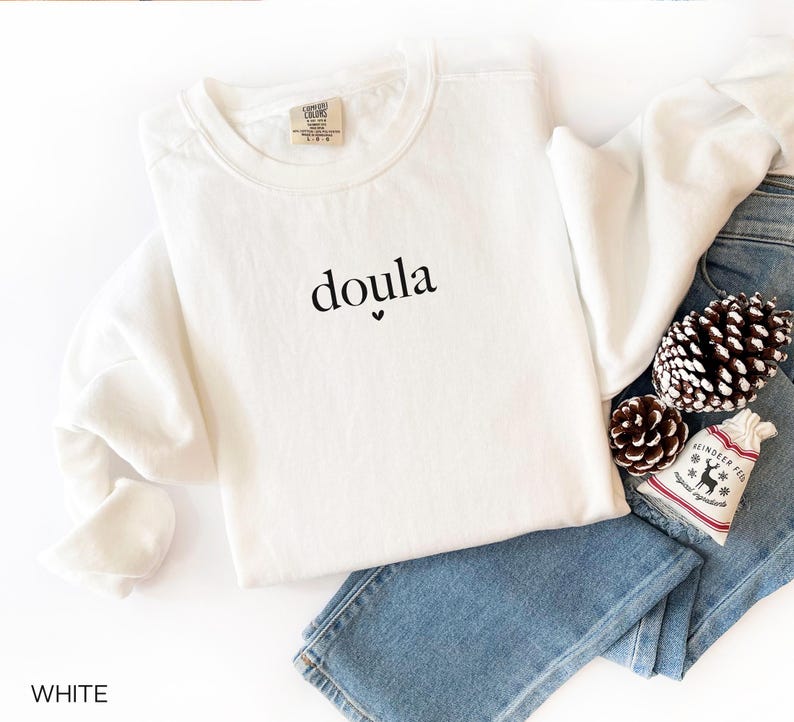 May include: White crew neck sweatshirt with the word "doula" printed in black. The sweatshirt is folded on top of blue jeans, with pinecones and a small decorative bag.
