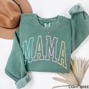 May include: Light green sweatshirt with the word "MAMA" in rainbow colors. The sweatshirt is a crew neck style with long sleeves.