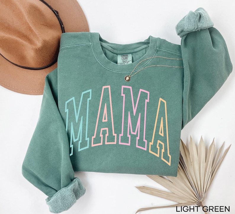 May include: Light green sweatshirt with the word "MAMA" in rainbow colors. The sweatshirt is a crew neck style with long sleeves.