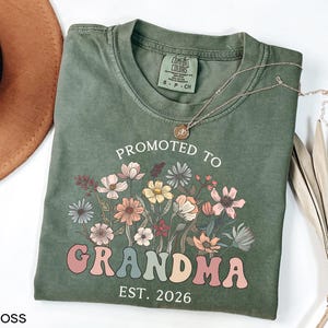 May include: A moss green t-shirt with the words "PROMOTED TO GRANDMA EST. 2026" in a retro font, accompanied by a floral design. A gold necklace rests on the shirt. The shirt is a Comfort Colors brand.