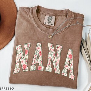 May include: Espresso-colored Comfort Colors t-shirt with the word "NANA" in a floral design. The letters are filled with pink roses, red berries, and green leaves. A silver necklace with a pendant is draped across the shirt.