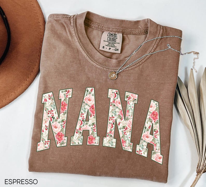 May include: Espresso-colored Comfort Colors t-shirt with the word "NANA" in a floral design. The letters are filled with pink roses, red berries, and green leaves. A silver necklace with a pendant is draped across the shirt.